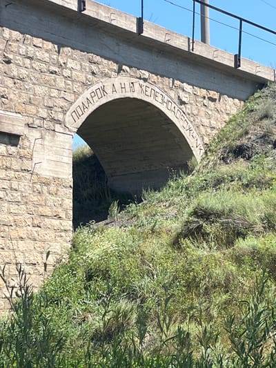 The image shows a stone bridge with an arch, a common feature in Aqtau, Kazakhstan. The arch has Cyrillic inscriptions that read "ПОДАРОК А Н.Ю. ЖЕЛЕЗНОДОРОЖНИК" which translates to "GIFT TO N.Y. RAILWAYMAN". The bridge is surrounded by overgrown green vegetation and a clear blue sky is visible above. The stone construction of the bridge suggests it is an older structure.