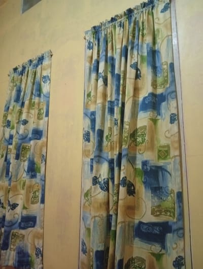 The image shows a close-up, eye-level view of two curtains hanging against a textured, light yellow wall. The curtains are made of fabric with a patterned design featuring abstract shapes, brushstrokes, and stylized leaves in shades of blue, green, and tan. The left curtain is slightly further away and appears narrower than the right curtain. The lighting suggests an indoor environment, possibly a room, with the light source likely coming from the front and slightly above. The wall has some imperfections and discoloration, indicating it is not perfectly smooth or recently painted. There are no people or animals visible in the image, and the focus is entirely on the curtains and the wall. The overall impression is of a simple domestic setting. The location is identified as Cihanjuang Rahayu, Indonesia.