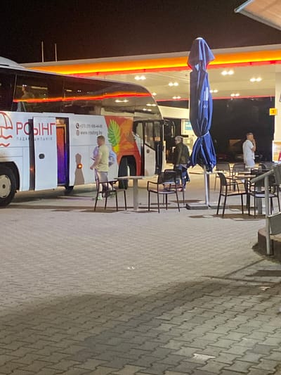 The image depicts a nighttime scene at a gas station in Horbów-Kolonia, Poland. A white bus with red lettering and graphics is parked in front of a gas pump island. The bus has its doors open, and several people are visible. One person, wearing a light yellow shirt and shorts, stands near the bus entrance. Another person, dressed in a dark jacket and jeans, stands to the left of the bus. To the right of the bus, a person in a white shirt and shorts is standing near the gas pumps. In the foreground, several tables and chairs are set up, suggesting an outdoor seating area. A dark blue umbrella, closed and covered, stands in the middle of the seating area. The gas station canopy is illuminated by bright orange and yellow lights. The ground is paved with interlocking gray pavers.
