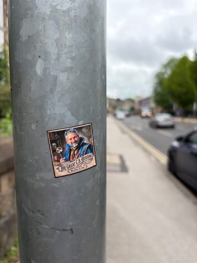 The image captures a close-up, angled view of a textured, galvanized metal pole, likely part of a street fixture. Dominating the lower-center of the frame is a rectangular sticker affixed to the pole. The sticker features a vintage-style illustration of an older man with white hair and a beard, wearing a blue and gold cloak, holding a crystal ball and surrounded by mystical paraphernalia like skulls and books.

Clearly visible text on the sticker reads: "THE GREAT C.B. HOUDINI" at the top right, and below that, "Mystifier & Seer." The sticker itself is slightly tilted within the frame. The pole shows signs of wear, with scuffs, scratches, and a mottled grey pattern characteristic of galvanized metal.

The background is heavily blurred and out of focus, showing an urban or suburban street scene under what appears to be an overcast sky. Faint shapes of buildings, cars, and green trees are discernible, suggesting daytime. The street appears to have vehicles, possibly parked or in motion, though no specific activity is clearly visible. Given the context, this scene is set in Harrogate, United Kingdom.