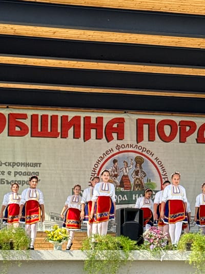 In Pordim, Bulgaria, a group of young girls in traditional Bulgarian folk costumes perform on a stage. The girls, with their hair adorned with floral wreaths, wear white blouses with intricate embroidery and red skirts with blue and gold trim. They are positioned across the stage, with some girls in the foreground and others behind them. A large banner with red Cyrillic lettering hangs behind them, partially obscured by the performers. The banner appears to say "ОБЩИНА ПОРОДИМ" (Municipality of Pordim) and "НАЦИОНАЛЕН ФОЛКЛОРЕН КОНКУРС" (National Folklore Competition). To the left of the banner, there is text in smaller font, and a circular emblem with depictions of traditional figures in folk attire. The stage itself is made of wood, with plants and a flower arrangement visible in the foreground. A black speaker is placed on the stage to the right of center. The backdrop suggests an outdoor event, with a wooden ceiling structure visible at the top of the image. The overall scene captures a cultural performance during a festival or competition in Pordim.