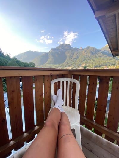 The image shows a person sitting on a white plastic chair on a wooden deck, with their legs crossed and facing the camera. The person is wearing white socks. A tattoo is visible on their right thigh. The deck overlooks a lush green mountain range under a clear blue sky with a few scattered clouds. The sun is shining brightly, indicating it is daytime, likely late morning or early afternoon given the angle of the sun. The mountains appear to be forested, with some buildings or structures visible nestled within the trees on the slopes. The wooden railing of the deck is prominent in the foreground. The overall scene conveys a sense of relaxation and appreciation of nature, with the person seemingly enjoying a peaceful moment outdoors. The location is identified as Lärchenwiese, Austria, which aligns with the mountainous landscape. No discernible text is visible in the image.