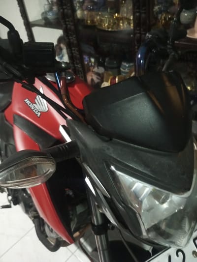 A stationary red and black Honda motorcycle is depicted indoors in the city of B. Nam Ngao, Laos. The motorcycle is positioned on a white tiled floor with dark grout, viewed from a front-quarter angle. Its matte red fuel tank bears the white "HONDA" logo and wing emblem. Black handlebars include a visible brake fluid reservoir on the left side and a blue metallic component centrally. The front cowl and headlight assembly are black, featuring clear headlight and right turn signal lenses. Chrome-colored front forks are partially visible, as is a white license plate with black characters at the bottom. The background, largely out of focus, reveals a dark, possibly wooden, mirrored cabinet displaying numerous reflective, metallic-toned decorative objects. No individuals or active interactions are visible.