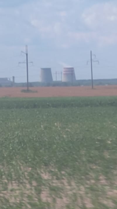 The image shows a wide shot of a rural landscape with industrial structures in the distance. In the foreground, a green field is visible, suggesting agricultural land. In the middle ground, there is a brown, barren strip of land. In the background, two large, cylindrical cooling towers, likely part of a power plant, are prominent. One of the cooling towers is emitting steam. Power lines and transmission towers are also present in the background, indicating the presence of an electrical grid. The sky is overcast with scattered clouds. The location appears to be in the vicinity of Красненскі сельскі Савет, Belarus, given the industrial elements juxtaposed with agricultural land, typical of such areas. The overall scene is one of an industrial facility situated within a rural environment.