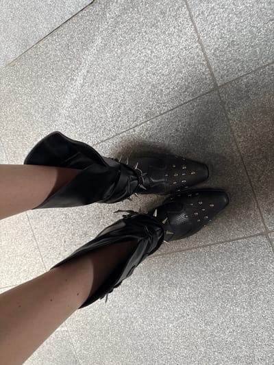 The image presents a first-person view of a person standing on a gray-tiled floor. The person is wearing black leather boots, adorned with metal studs on the front and spikes around the ankle. The boots have a pointed toe and an open, flared design at the top, revealing the person's legs. The floor appears to be indoors, possibly in a public building or commercial space, given the tile pattern. The location is not provided within the image and would require additional contextual information.