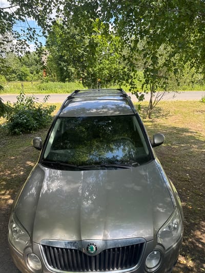 This is a high-angle, medium shot of the front of a silver Skoda Yeti parked outdoors. The car is angled slightly towards the right side of the frame. The hood of the car is visible, along with the grille, headlights, and windshield, which reflects the surrounding trees and sky. The car has a roof rack. The background consists of green trees, grass, and a road with a power line pole visible in the distance. The scene appears to be in a rural or suburban area. The lighting suggests it is daytime, with sunlight filtering through the trees and creating shadows on the car and the ground. The location is specified as Мазалаўскі сельскі Савет, Belarus. There are no people or animals present in the image.