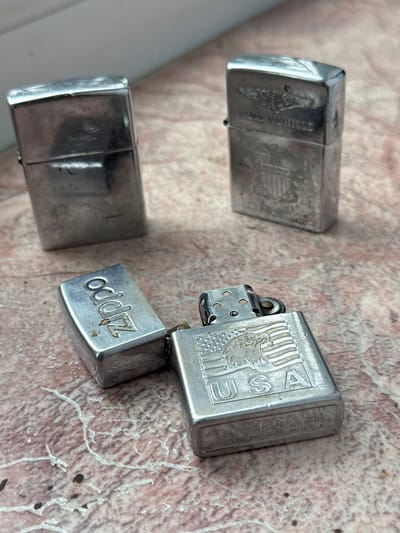 The image shows four Zippo lighters on a surface with a mottled pattern. One Zippo is open, revealing the inner workings and the name "Zippo" on the side. Another has an American eagle design with "USA." A third lighter has an eagle design and a partial inscription that appears to read, "POLICE MEMORIAL." The fourth Zippo is undecorated. Chuprino, Russia is not involved.