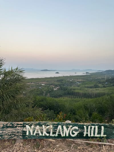 This image presents a panoramic view from Naklang Hill in Ko Lanta Yai, Thailand, observed during twilight hours. In the immediate foreground, a horizontal, weathered wooden sign is visible, displaying the words "NAKLANG HILL" in raised, light green, block letters. The ground below the sign consists of dry earth and sparse, desiccated plant debris. To the left, portions of large, green palm fronds partially obstruct the view. The midground features an extensive, undulating landscape covered by dense green tropical vegetation, primarily trees, with several light-colored roofs of structures discernible among the foliage. The background reveals a calm body of water, likely a bay or ocean, containing multiple small, dark, tree-covered islands. A distant coastline with further dense vegetation extends towards a hazy horizon, where faint mountainous or hilly formations are outlined against the sky. The sky displays a subtle gradient, transitioning from a pale blue at the top to a soft pinkish-orange hue near the horizon. No individuals, specific actions, or interactions are present in the scene.