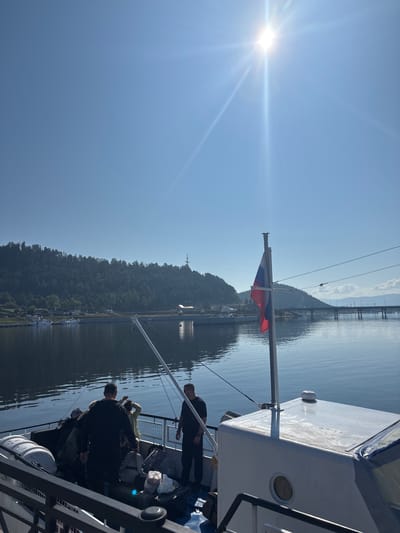 This is a vertical, eye-level shot of a bright, sunny day on a body of water, possibly a lake, in Turka, Russia. In the foreground, the deck of a white boat is visible, with several people gathered. A Russian flag flies on a pole to the right. The water is calm and reflects the clear blue sky. Across the water, a lush green, tree-covered hill rises. In the distance, a bridge spans the water, and buildings and infrastructure are visible along the shoreline. The sun is high in the sky, casting bright rays and lens flares across the image.