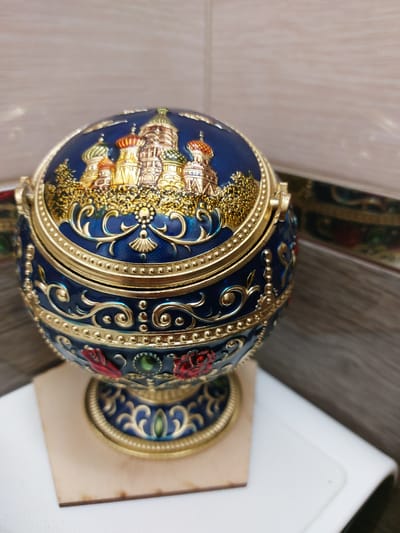 A close-up, eye-level shot shows an ornate, dark blue, egg-shaped decorative box with gold detailing and a lid. The lid and upper portion of the box are adorned with a raised, golden depiction of what appears to be St. Basil's Cathedral in Moscow, Russia, characterized by its multi-colored onion domes and intricate architectural elements. The lower part of the box features scrolling gold patterns, red rose embellishments, and green jeweled accents. The box is resting on a small, light-colored wooden square, which is placed on a white surface. The background consists of a textured wall with horizontal bands of light and dark tones, possibly wood paneling or textured wallpaper. The image was taken indoors, and the lighting suggests a bright, possibly natural, light source illuminating the subject. There is no indication of the location being specifically in Nakhabino, Russia; it is a general decorative object with Russian architectural motifs.