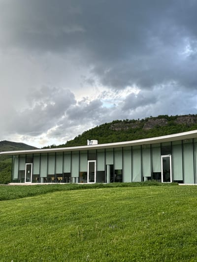The image shows a building with a curved facade and large windows, adjacent to a grassy area. Behind the building, there is a cliff face with green vegetation. The sky is filled with dark, dramatic clouds. The building's interior appears to have tables and chairs. The scene is most likely located in Chkalov, Armenia, given the prompt.