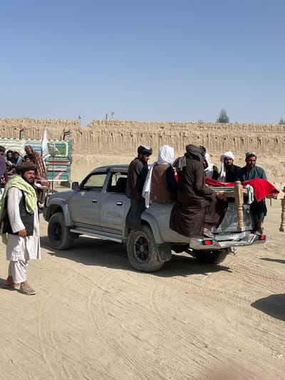The image depicts an outdoor scene in Spin Boldak, Afghanistan, under a clear, bright sky. A silver-grey Toyota pickup truck, modified with aftermarket wheels and off-road tires, occupies the central mid-ground. In the bed of the truck, approximately five men, dressed in traditional attire including shalwar kameez and various head coverings, are gathered. One man, wearing dark brown, sits on a wooden frame within the truck bed, which is draped with a prominent red fabric, suggesting it carries an object or person. Another man in dark clothing is visible partially standing on the truck's running board. To the immediate left of the truck in the foreground, a man with a dark beard, wearing a white shalwar kameez, a dark vest, and a light green scarf, stands with his body angled left and head turned towards the scene, wearing open-toed sandals. In the left background, a larger group of men is visible near a multi-colored truck or lorry, painted in green and blue, featuring a white flag possibly affixed to its side. The ground is unpaved, dusty, and light brown, showing numerous tire tracks. Stretching across the entire mid-background is a tall, textured mud or adobe wall structure, appearing weathered with a noticeable breach or opening near its center.