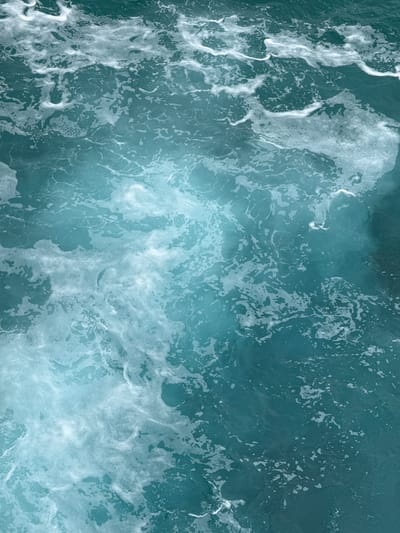 This is an aerial, close-up photograph of churning, foamy ocean water. The water is a deep teal color with lighter, swirling patterns of white foam breaking on the surface. The foam creates intricate, organic shapes, resembling delicate lace or brushstrokes across the darker water. Some areas of the foam are thick and opaque, while others are thin and translucent, allowing the blue of the water to show through. The texture of the water is dynamic, with a sense of movement and turbulence suggested by the swirling patterns and breaking waves. There are no visible people, objects, text, or clear time of day or weather cues, beyond the implied presence of the sea. The image captures the raw, elemental beauty of the ocean's surface in motion, likely somewhere along the coast of Llanes, Spain, known for its dramatic coastlines and active waters. The overall mood is one of natural power and serene abstraction.