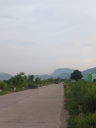 A moderately wide, paved road stretches into the distance, bordered by lush green vegetation on both sides. In the foreground, the road surface shows signs of wear, with some cracks and debris. White cylindrical markers with red tops are spaced along the right edge of the road.
Several vehicles are visible on the road. A truck is moving away from the viewer in the middle distance. To the right of the truck, a person is riding a motorcycle. Further down the road, another motorcycle is visible. Closer to the left side of the road, a two-wheeled cart loaded with foliage is parked, and a person is standing next to it. A person on a motorcycle is also seen in the distance on the right side of the road.
The landscape features rolling hills and mountains in the background under a hazy, overcast sky. On the right side of the road, a modern, multi-story building with green and white facades is visible among the trees. Dense, tall green grass lines the right edge of the road. The overall impression is a rural or semi-rural setting in Vietnam.