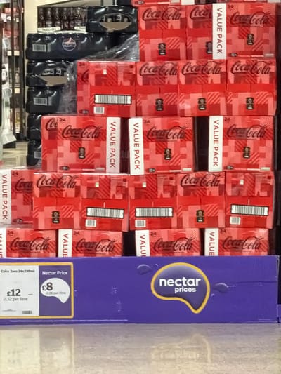 This image depicts a supermarket shelf stocked with Coca-Cola "Value Pack" 24-can cartons. The cartons are red with white "VALUE PACK" lettering and Coca-Cola branding. To the right of the Coca-Cola cartons is a blue "Nectar Prices" sign advertising "Coke Zero 24x330ml" for £8 (£1.52 per litre) as a Nectar Price with a standard price of £12. To the left, partly visible, are other products on the shelf, including some black packaged goods. Based on the "Nectar Prices" logo, the location is likely a Sainsbury's supermarket in Pity Me, United Kingdom, though this can't be conclusively determined from the image alone.