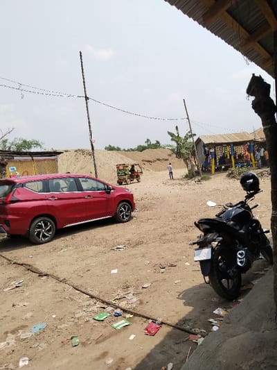 The image shows a dirt road in front of a small shop in Pangasia, Bangladesh, located at coordinates (latitude: 22.705, longitude: 90.565). A red SUV is parked on the left side of the road, facing the camera. On the right side, a black motorcycle is parked, facing the same direction as the SUV. A small auto rickshaw can be seen further down the road. A few people are visible in the background, walking around or standing near the shop. There are several power lines strung across the sky, and the ground is covered with scattered litter. The shop is a small, single-story building with a corrugated metal roof and a few windows. There is a pile of sand in the background, which is likely used for construction. The image was taken on a clear, sunny day. The scene is typical of a rural or semi-rural area in Bangladesh.