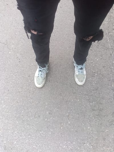 The image shows a person standing on an asphalt surface in Park Hill, United States. The person is wearing black ripped jeans and light-colored sneakers with blue laces. The jeans have large tears at the knees. The asphalt surface is cracked and shows signs of wear.