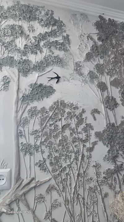 This is a close-up, eye-level photograph of a textured wall mural depicting a forest scene. The mural is rendered in shades of grey and white, with a highly tactile, bas-relief style. Prominently featured are the trunks and branches of several trees, some with dense, textured foliage, and others with more sparse, delicate branches. In the upper center of the image, a small, dark silhouette of a bird in flight, likely a swallow, is positioned against the lighter background. The lower portion of the mural transitions to smaller, thinner plants and grasses, also rendered with significant texture. A white electrical outlet is visible on the left side of the frame, at the base of a large tree trunk. The overall impression is one of naturalistic detail and artistic craftsmanship. The photograph's metadata indicates it was taken in Nosy, Ukraine.