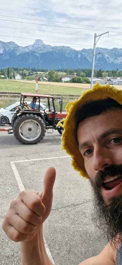 A man with a beard, wearing a yellow fringed hat, is taking a selfie. He is looking at the camera with a surprised or excited expression, mouth open. In the background, a red tractor with a driver is visible, parked next to a white car. The tractor is on what appears to be a road or parking lot with painted lines. Beyond the road, there are fields of crops, houses, and a mountain range under a cloudy sky. A power line structure is also visible in the distance. The scene suggests a rural or semi-rural setting, likely in Uetendorf, Switzerland, during the daytime. The overall mood appears to be casual and possibly adventurous.