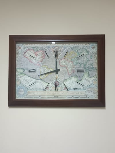 A wall clock with a map of the world is mounted on a beige wall. The clock has a dark brown frame and Roman numerals indicating the hours. The map on the clock face is detailed, showing continents, oceans, and various geographical features. The clock hands are black. The overall scene is static and shows no people or any activity. It is impossible to determine the location in Luanda, Angola from the provided image.