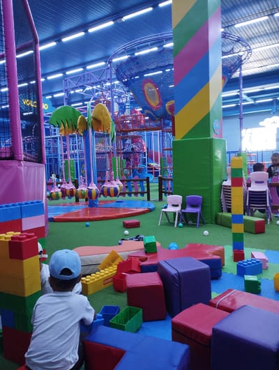 The image depicts an indoor children's play area. In the foreground, a young child wearing a white t-shirt and a blue baseball cap is playing with large, colorful building blocks. To the left, a stack of large, red, yellow, and blue blocks are visible, resembling oversized Lego bricks. In the background, various play structures are present, including netting, colorful slides, and hanging elements that appear to be part of a soft play or adventure zone. The flooring is covered in green artificial turf. Several small, colorful chairs are placed in the mid-ground. The overall environment is brightly lit with overhead fluorescent lighting and features a vibrant, multi-colored striped pillar in the mid-right. The scene appears to be located in Talas City, Kyrgyzstan, an indoor family entertainment center.