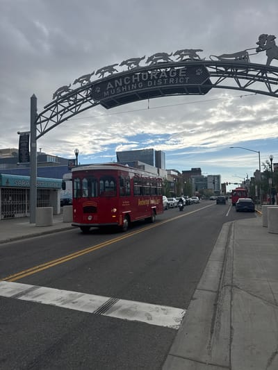 The image shows a street scene in Anchorage, Alaska. A red trolley bus with "Anchorage Trolley Tours" written on its side is stopped at the side of the road. The trolley has multiple windows and is adorned with ornate metalwork. A large metal archway with the words "ANCHORAGE MUSHING DISTRICT" is visible to the left of the trolley. The archway features a silhouette of sled dogs pulling a sled. The street is paved and has yellow double lines dividing the lanes. Other vehicles, including cars and a motorcycle, are seen on the road. Buildings line the street, and the sky is partially cloudy, suggesting it is daytime. The overall scene depicts a tourist-oriented area in an urban setting. Visible text also includes "Santos" on a building and "SARAH GIFT CACHE" on a storefront.