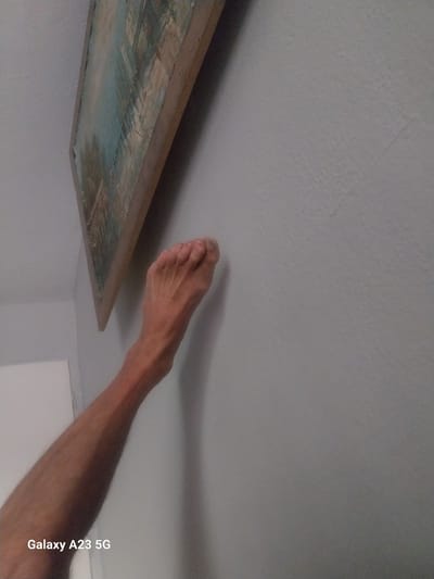 An overhead shot shows a human leg and foot pressed against a textured, light gray wall. The leg extends from the bottom left corner of the frame upwards and to the right, its skin toned brown with visible hair. The foot, with its toes splayed, is pressed flat against the wall. Above and to the left of the leg, a framed painting hangs on the wall. The painting is rectangular, with a wooden frame, and its contents appear to be an abstract landscape in shades of blue, white, and brown. A watermark in the bottom left corner reads "Galaxy A23 5G." The scene is lit from above and to the right, casting a subtle shadow from the leg onto the wall. The location is not discernible from the image.
