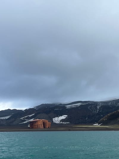 A landscape with a cloudy sky dominating the upper left. A dark, rocky mountain with patches of snow runs vertically down the right side of the image. At the base of the mountain, near the water, stands a distinctive copper-colored building. The water is a muted turquoise color and occupies a narrow strip along the right edge.