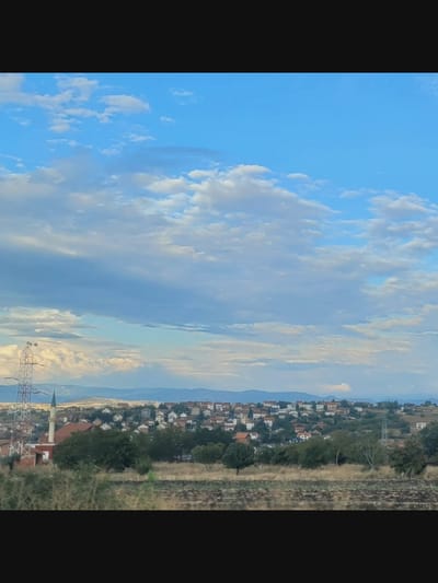 The image displays a vast, open sky filled with scattered white and gray clouds against a vibrant blue backdrop. Beneath the sky, a sprawling residential area is visible, characterized by numerous houses with red-tiled roofs, clustered together on undulating terrain. A prominent minaret, likely belonging to a mosque, stands tall amidst the buildings, alongside a tall, lattice-like power transmission tower. The foreground consists of a field of dry grass and vegetation, suggesting a rural or semi-rural environment. The overall scene conveys a sense of a settled community under a dynamic sky.