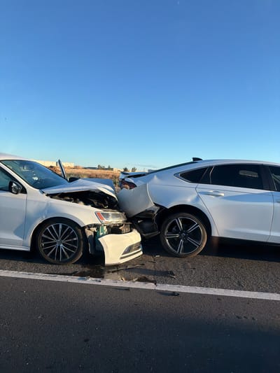 Two white cars have been involved in a collision, with the front of the lower car severely damaged and the rear of the upper car also showing significant damage. The cars are positioned on a paved road, likely a highway, indicated by the clear lane marking and the presence of roadside vegetation in the background. The sky is a clear, bright blue, suggesting it is daytime with good weather. The sunlight casts shadows, indicating the sun is relatively high in the sky. The scene appears to be in Sacramento, United States, given the provided location context. No people are visible in the image, and there is no visible text. The damaged condition of the vehicles suggests an accident has recently occurred.