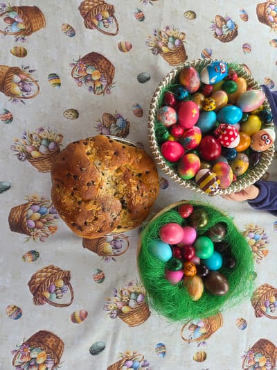 The image shows a table covered with a patterned tablecloth depicting Easter baskets and eggs. On the table are two baskets filled with colorful Easter eggs, one of which is held by a person with their arms visible, and a round Easter bread (Kozunak) with almonds and chocolate chips. The scene is likely set indoors, potentially in a home setting, but there is no direct evidence to link it to Tompsan, Bulgaria.