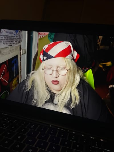 The image displays a screen showing a person, with a laptop keyboard partially visible in the foreground to the right. The screen is oriented sideways.
The person on the screen has pale skin, long, light blonde hair, and is wearing round, wire-frame glasses and red lipstick. A bandana with a prominent red and white stripe pattern, resembling a section of the US flag, covers part of their head. They are dressed in dark clothing, with a flash of bright neon green or yellow fabric visible around their neck area. The person's eyes are partially closed or cast downwards, giving them a pensive or serene expression.
The background behind the person on the screen is dark and indistinct. Below the displayed person, on what appears to be a wall or surface within the displayed content, are posters or papers. Partially visible text on these items includes "Deat" and "AMERICANA NDEPENDENTE DE".
To the right, the black keys of a laptop keyboard are visible, along with parts of the laptop's bezel. Timecodes "01:53:01" and "-00:14:16" are visible on the screen's edge, suggesting the display shows a video. The overall lighting of the physical environment where the photograph was taken is dim, with the screen providing the main illumination. The location context for the image is Torrent, Spain, although no specific visual cues in the image confirm this.