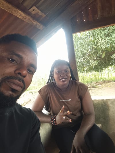 Two people are in a selfie, a man on the left and a woman on the right. The man is closer to the camera, with his face and the left side of his body visible. He has a beard and is wearing a black t-shirt. The woman is seated to his right, facing the camera and smiling. She has braided hair and is wearing a brown t-shirt with text on it. She is also wearing bracelets and rings. They are under a wooden and corrugated metal roof structure. Behind them, there is vegetation, including a large tree with green leaves and a field of crops. The overall setting appears to be outdoors, possibly a rural area in Adikpo, Nigeria. The lighting suggests it is daytime. The woman's expression and the selfie format suggest a casual and friendly interaction. The text on the woman's shirt reads "RELENTLESS BUSINESS".