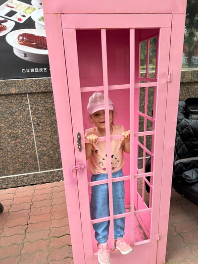 A young girl, wearing a pink baseball cap, a peach-colored t-shirt with cartoon faces, and light blue pants, stands inside a bright pink telephone booth. She is smiling and holding onto the bars of the booth. The booth is open on one side, revealing the interior. In the background, to the left of the booth, a black, textured wall is visible. To the right of the booth, a black quilted jacket or bag is draped over a surface. The ground in front of the booth is paved with red interlocking bricks. A sign with Chinese characters, likely indicating a restaurant or establishment, is partially visible on the left side of the frame, suggesting the location is in the city of Hunchun, China.