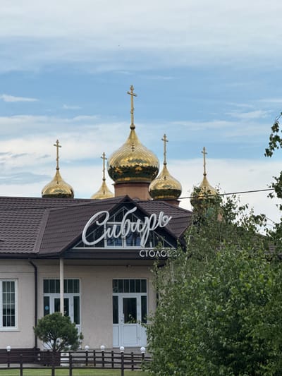The image depicts a building with multiple golden domes, characteristic of Russian Orthodox churches, topped with crosses. The building's roof is dark brown, and the facade is light-colored with white-framed windows and doors. A white, illuminated sign in cursive script is mounted above the entrance, with smaller text below. Lush green trees and foliage are visible in the foreground and partially obscure parts of the building. The sky is overcast with clouds. The location is identified as being in the city of Shushenskoye, Russia.