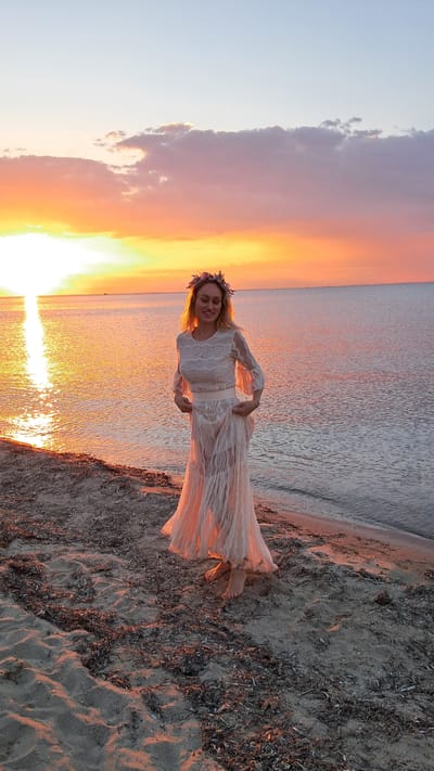 A woman stands on a sandy beach at sunset, wearing a long, light-colored dress and a floral crown. She is facing forward, smiling slightly, with her hands on her dress. The sun is setting over the calm sea, casting a warm, orange glow across the sky and water. The beach is covered in sand and scattered debris. The location is Agia Triada, Greece.