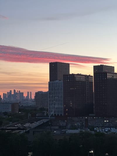 The image displays a panoramic view of an urban landscape at either sunrise or sunset, observed from an elevated vantage point. The scene is dominated by a dramatically colored sky and the silhouettes of city buildings.
The upper portion of the frame features a sky that transitions from a light, pale blue-grey at the top left to vibrant reds and oranges along a long, horizontal cloud formation stretching across the mid-section. Below this striking cloud band, the sky softens into warm yellow and peach tones closer to the horizon. A faint, thin line, possibly an aircraft contrail, is visible in the upper left sky.
In the foreground and midground, several large, block-like multistory buildings stand in dark silhouette against the luminous sky. These structures appear to be apartment blocks or commercial buildings, characterized by repetitive grid-like window patterns. Scattered lights within some windows indicate artificial illumination, suggesting the time is dusk or dawn. To the right, dark green tree foliage partially obscures views of lower structures.
Further in the distance, a sprawling city skyline extends towards the horizon, also appearing in silhouette. Numerous smaller buildings and the faint glimmer of distant city lights contribute to the impression of a vast urban environment. There are no people visible, nor is there any discernible text or signage in the image. The media captures a tranquil moment over a bustling city, consistent with an urban setting in Ukraine.