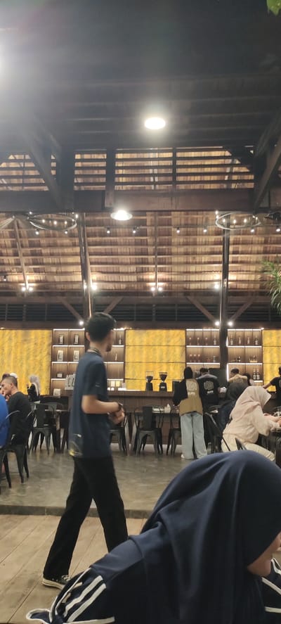 This image captures an indoor scene, likely a cafe or restaurant, with a warm, rustic ambiance due to its wooden ceiling and exposed beams. The lighting suggests it is evening or nighttime, with several circular overhead lights illuminating the space.
In the foreground, a person wearing a dark blue t-shirt and black pants walks from left to right, facing away from the camera. They are wearing black sneakers. To the right of this person, another individual, also wearing a dark blue hooded jacket with white stripes on the sleeves and a dark blue hijab, is partially visible, looking towards the right.
The mid-ground reveals a bar area with shelves stocked with bottles and other items. Two large coffee grinders are visible on the counter. Several tables and chairs are arranged throughout the space, with some people seated. One person in the background is wearing a light brown hooded jacket over a black top, and another is wearing a black jacket with the word "HEAVEN" printed on it. Further back, a person in a light pink hijab and white pants is seated. The overall atmosphere appears relaxed, with people socializing or enjoying their time.
The walls behind the bar area are decorated with a textured, golden-yellow fabric or material. The floor is a combination of concrete and wooden planks. The architecture of the ceiling, with its layered wooden slats, is a prominent feature. The setting evokes a cozy, communal gathering place. There is no visible weather information or overt emotional cues, but the dim lighting and presence of patrons suggest a comfortable, possibly late-evening ambiance. The text "HEAVEN" is visible on the back of a jacket in the background.