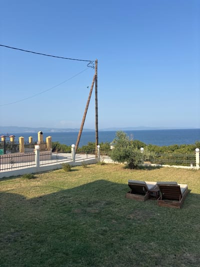 This is a vertical, slightly tilted, outdoor, daylight photograph taken in Nea Moudania, Greece. The scene captures a panoramic view of the sea and sky, with a grassy lawn in the foreground. The sky is a clear, bright blue, suggesting a sunny day.
Dominating the left side of the image is a wooden utility pole with wires extending from it. The pole is angled, running from the bottom left corner upwards towards the top center.
The foreground features a neatly mowed green lawn that slopes downwards. Along the edge of the lawn, a paved walkway runs parallel to the sea. This walkway is bordered by a black metal fence with decorative elements. Beyond the fence, there are several clusters of what appear to be outdoor seating arrangements. These seating structures are built with concrete bases and wooden slatted tops, some of which are open and appear to be designed to hold cushions. White globe lights are visible along the walkway, illuminating the area.
In the background, the deep blue sea stretches out to the horizon, meeting a hazy blue sky. The distant shoreline is visible as a faint line of land. The overall impression is one of a serene and pleasant coastal setting, possibly a resort or a residential area with a view of the sea. No people are visible in the image. The time of day is clearly daylight, with strong shadows cast by the seating structures onto the lawn, indicating the sun is high in the sky.