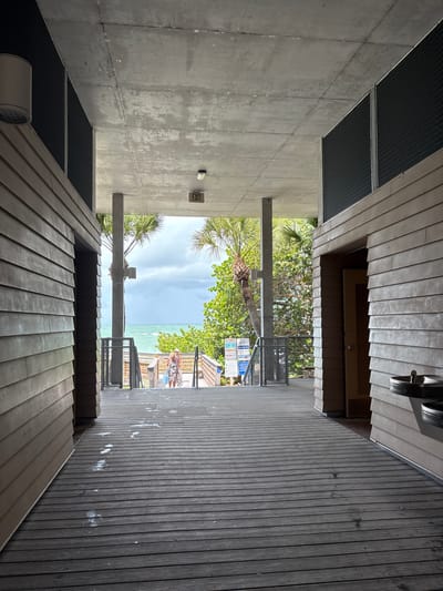 The image depicts a wooden boardwalk leading towards a beach access point. On the left and right sides of the boardwalk are wooden structures, possibly restrooms or changing facilities, with siding. In the distance, the ocean is visible under a cloudy sky, with palm trees and lush green foliage framing the view. Two individuals, a woman and a child, are walking up a set of wooden stairs towards the beach access. The boardwalk is wet in places, indicating recent moisture. An "EXIT" sign is visible on the ceiling of the structure. The scene appears to be a public beach access in Englewood, United States.