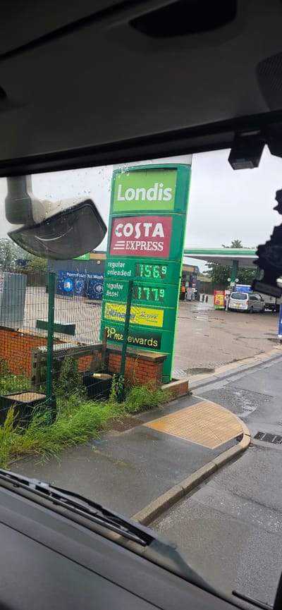 The image depicts a gas station sign with fuel prices displayed. The sign is green and has several panels. The top panel reads "Londis" in white text. Below that, a red panel displays "COSTA EXPRESS". Further down, a green panel shows "regular unleaded" with a price of "156.9" and another panel shows "regular diesel" with a price of "177.9". At the bottom of the sign, a green panel reads "3p me ewards".
In the background, there is a canopy over the gas pumps. There are several vehicles, including cars and vans, at the station. There are also some individuals, possibly gas station employees, wearing orange vests. To the left of the sign, there is a car wash facility.
The ground is wet, indicating it may have rained recently. The scene appears to be set in an outdoor environment, likely a convenience store or gas station. The image is taken from inside a vehicle, with a windshield wiper visible in the foreground. The location is identified as Tameside, United Kingdom.