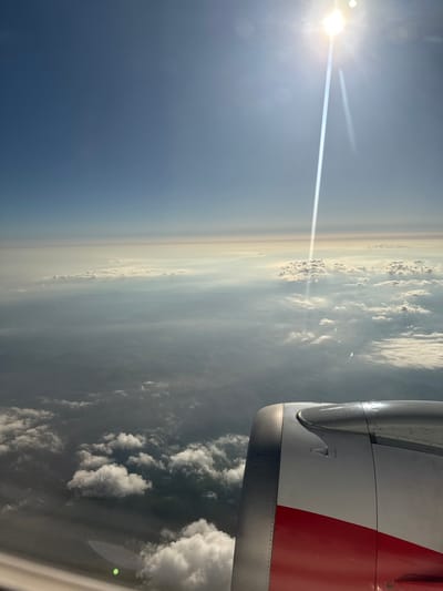 This is an aerial photograph taken from an airplane window, looking out at the sky and clouds. The sun is visible in the upper right quadrant of the image, creating a bright lens flare that streaks down towards the horizon. Below the sun, layers of clouds are present, with some appearing wispy and others more dense. The landscape below the clouds is hazy and appears mountainous, with faint outlines of hills and valleys. In the foreground, a portion of the airplane's engine nacelle is visible, featuring a metallic silver upper section and a red stripe on the lower section. The scene suggests an airplane in flight over a terrestrial landscape, likely during daytime. The location of Brakyovtsi, Bulgaria is not visually evident in this image.