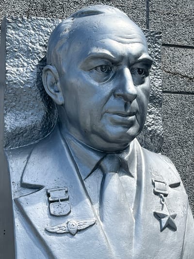 A silver statue of a man's head and upper torso is depicted in close-up, angled from above and to the left. The statue is made of a reflective material, with shadows creating depth and highlighting the sculpted features. The man has closed eyes and a partially open mouth, suggesting a state of rest or perhaps death. He wears a uniform with visible insignia, including a star on his left shoulder and what appears to be a medal or badge on his right chest. The texture of the statue's surface varies, with smoother areas on the face and clothing contrasting with a rougher, almost granular texture on the base and background.

The background is composed of two distinct textures: a dark, granular surface that resembles rough stone or concrete, and a lighter, crinkled metallic surface beneath the statue's head. The overall color palette is monochromatic, dominated by silver, grey, and dark tones, with the silver of the statue reflecting light. The lighting suggests it is daytime, possibly sunny, given the sharp contrasts and highlights. The location is identified as Takhtamukai, Russia, though no specific landmarks are visible within the image itself to confirm this. The statue appears to be a memorial or monument.