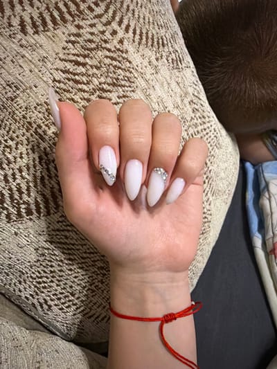 A woman's hand with almond-shaped white acrylic nails adorned with small rhinestones is resting on a textured, neutral-colored cushion. The woman is also wearing a thin red string bracelet on her wrist. In the background, a person's head with short dark hair is visible, along with a folded blue and white patterned cloth. The scene appears to be indoors, on a couch. The location is Zmeitsa, Bulgaria.