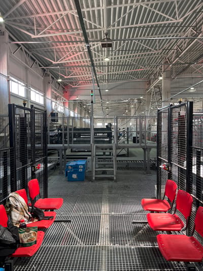 The image shows an interior view of a large, industrial building, possibly a warehouse or factory, located in Nerubais'ke, Ukraine. The space is characterized by its metal grating flooring and an overhead network of white metal beams and corrugated roofing. Various structures, including elevated platforms, staircases, and conveyor systems, are visible throughout the building.

In the foreground, a row of four bright red plastic chairs is situated on the metal grating. One chair has a plaid blanket and a black bag draped over it. Another chair has a package of "CRAFT" cookies and a bottle of dark liquid placed on it. The chairs are positioned on a raised platform behind a black metal fence.

Further into the building, a blue plastic crate is placed on the floor. Several other elevated platforms with stairs and railings are present, suggesting a multi-level operational area. The overall environment appears to be in a state of operation or preparation for use, with lighting fixtures and industrial equipment visible. There are no people in the scene.