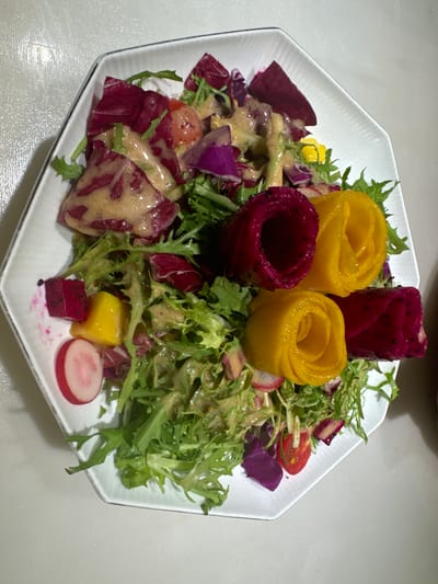 A vibrant and artfully arranged salad is presented on a white octagonal plate, set against a plain light grey surface. The dish features a colorful assortment of fresh ingredients, meticulously placed for visual appeal, consistent with a dining experience in Fangshan District, China.

The base of the salad consists of a mix of leafy greens, including frisée or endive with its characteristic curly leaves, and possibly darker red lettuce or radicchio. A creamy, speckled dressing, light brown or tan in color, is generously drizzled over the greens and other components.

Central to the presentation are four prominent "roses" made from fruit: two bright yellow ones, likely carved from mango, and two deep magenta ones, crafted from red dragon fruit, showcasing its distinctive tiny black seeds. These decorative elements are nestled among the greens.

Other visible salad components include slices of radish with red skin and white flesh, small cubes of yellow mango and red dragon fruit, pieces of red cabbage or radicchio, and halved cherry tomatoes. There are also larger, dark red pieces, possibly slices of beetroot or another type of red root vegetable, and smaller purple elements mixed throughout. A tiny yellow edible flower or fruit piece is also visible among the greens.

The image is a close-up, focusing entirely on the salad, suggesting an indoor setting with artificial lighting. There are no people visible, nor any text. The overall impression is one of a fresh, healthy, and aesthetically pleasing culinary creation.