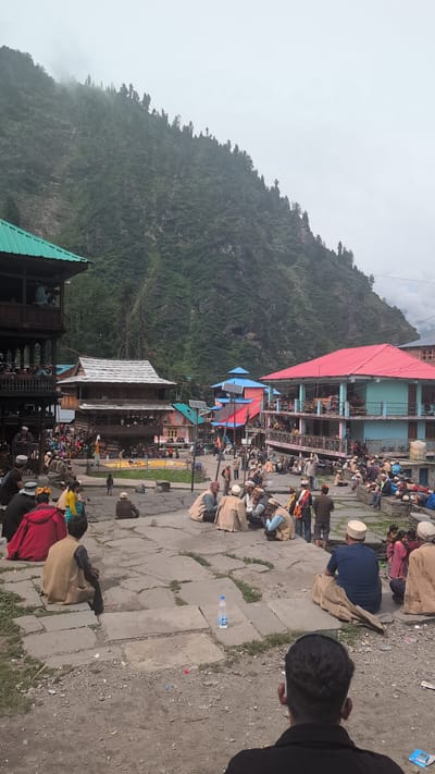 The image captures a lively scene in the village of Malana, India, nestled amidst a dramatic mountain landscape. The architecture is distinctly traditional, with wooden structures featuring multi-tiered balconies and sloping roofs, some adorned with vibrant red and blue hues. People, mostly men, are gathered in the central open area, dressed in traditional attire, many wearing distinctive woolen caps. They are seen sitting, conversing, and observing. Some are seated on the stone paving, while others occupy raised platforms. In the background, a steep, verdant mountain slope rises, partially shrouded in mist, indicating a high-altitude location. The atmosphere appears to be that of a community gathering or a local event. The ground is paved with large, irregular stone slabs, with patches of dirt and sparse vegetation. Various everyday objects are visible, including a water bottle placed on the ground and some scattered debris. The overall impression is one of a cultural snapshot of life in a remote Indian mountain village.