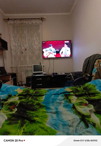 This media file shows an indoor scene, likely a bedroom in Plesetsk, Russia, at night. The primary focus is a bed covered with a brightly colored duvet featuring a nature-inspired pattern of trees and water. In the background, a television screen displays two men; one is playing a guitar, suggesting a musical performance or video is being watched. To the left of the television, a computer monitor and a desktop tower are visible, indicating a home office setup or a general use area. A curtain with a floral pattern hangs on the left side of the frame, partially obscuring a window, and suggests a domestic setting. A dark entertainment stand holds the television and computer equipment. Visible text includes "23:01" and "2304 20" on the television screen and a digital clock, respectively, confirming the late hour. The bottom of the image displays camera information: "CAMON 20 Pro" and "24mm f/1.7 1/33s ISO1112." The overall impression is a cozy, lived-in space at rest, with entertainment and technology present.