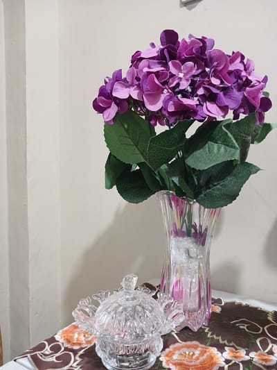 A close-up, eye-level shot captures a still life arrangement on a table in Geudonggeudong, Indonesia. The background is a plain, cream-colored wall with vertical shadows suggesting uneven lighting. In the foreground, a decorative brown table runner with embroidered orange and white floral patterns lies partially visible. To the left, a clear, intricately cut crystal bowl with a lid sits on the runner. To the right, a tall, clear glass vase with subtle pink and purple striations holds a bouquet of artificial purple hydrangeas with green leaves. The vase contains clear decorative crystals at its base.