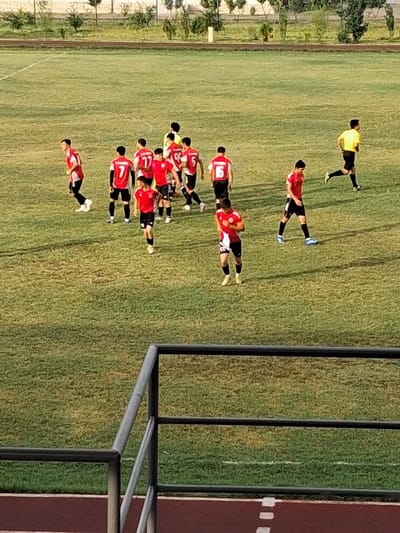 In Bustan, Uzbekistan, a group of young men, likely a soccer team, are on a grassy field. The team is wearing red and black uniforms, with one player in a yellow shirt. The field appears to be a soccer pitch, with white lines marking the boundaries. The players are scattered across the field, some walking and others appearing to be in motion. In the background, there are trees and a fence, suggesting an outdoor sports facility. The overall scene depicts a typical moment during a soccer match or practice.